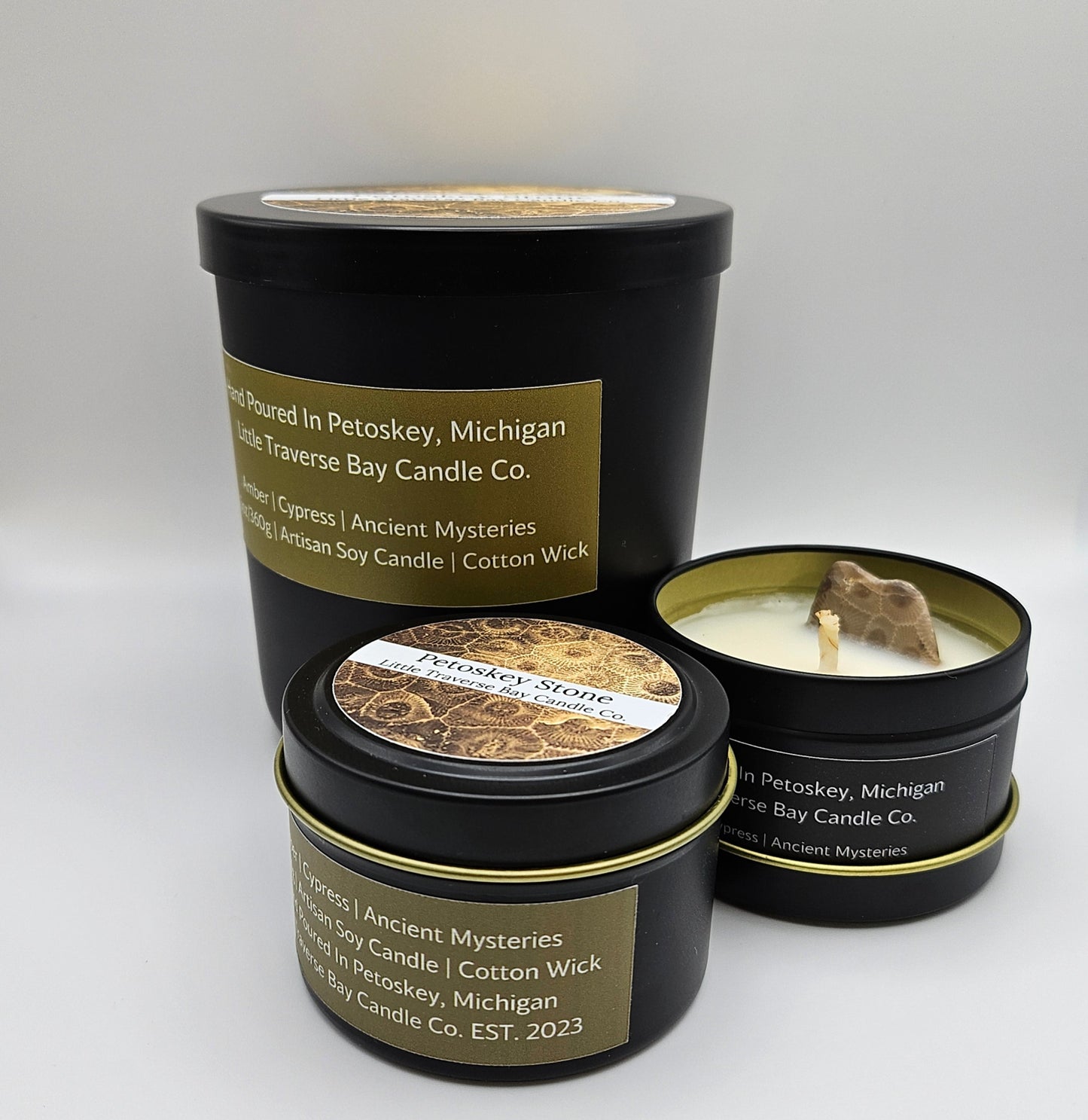 Three candles sit flat on a white background. The back candle is the largest with 12.5 ounces (oz) and the front two (2) have 4 oz of candle contents. The front small candle is closed with the top lid on. The other small candle is open and shows a cotton wick with a Petoskey stone popping up embedded in the wax. The large candle is also closed with a lid.