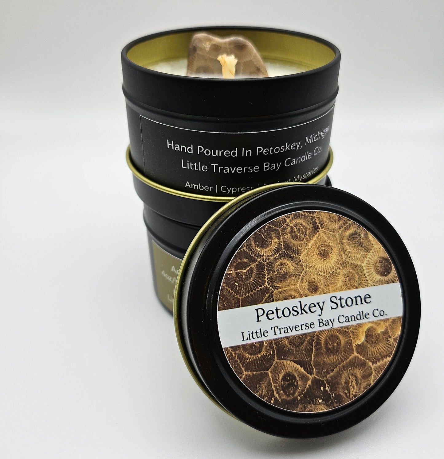 Two (2) 4 ounce soy candles in black tins stacked atop each other on a white background. The lid to the top candle is off and leaning against them with the top label showing. They say Petoskey Stone by Little Traverse Bay Candle Co. A Petoskey stone peeks up from inside of the top candle sitting behind a cotton wick.