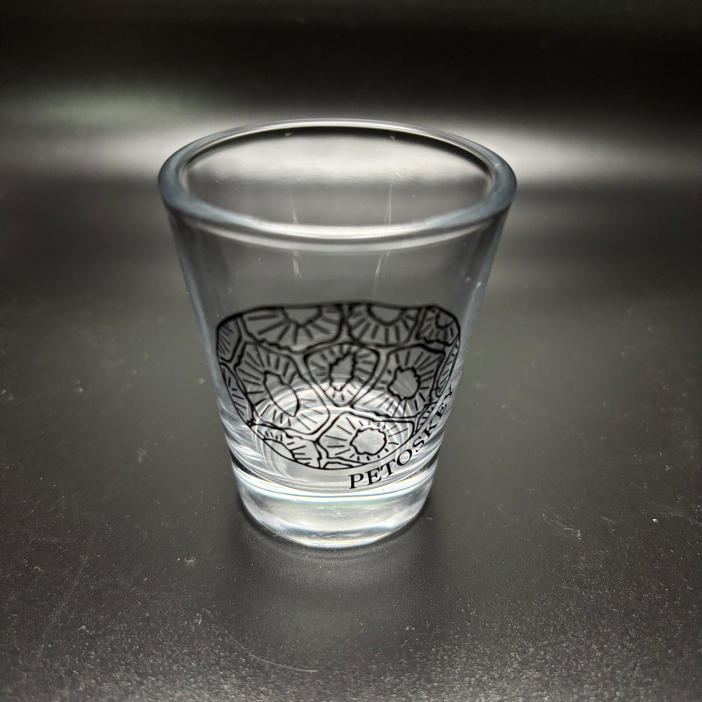 Clear glass shot glass with black design of a petoskey stone and text "Petoskey" on a dark surface