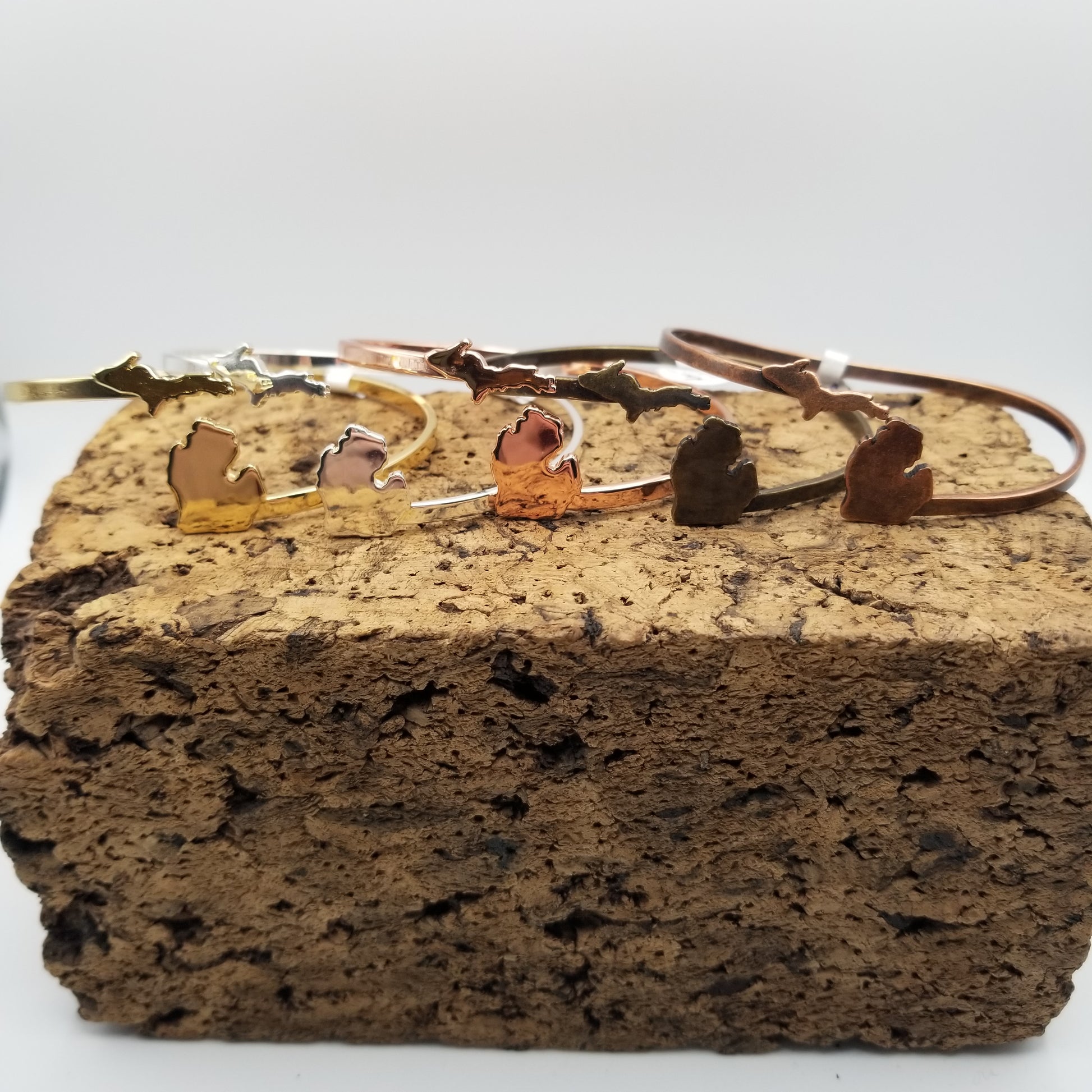 Line up of five wrap around adjustable cuffs on top of a block of cork. Each bracelet has the upper peninsula of michigan on the left side and the lower peninsula on the right. They are gold, silver, copper, antiqued silver, and antiqued copper, respectively.