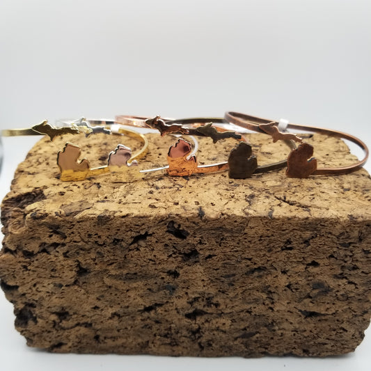 Line up of five wrap around adjustable cuffs on top of a block of cork. Each bracelet has the upper peninsula of michigan on the left side and the lower peninsula on the right. They are gold, silver, copper, antiqued silver, and antiqued copper, respectively.