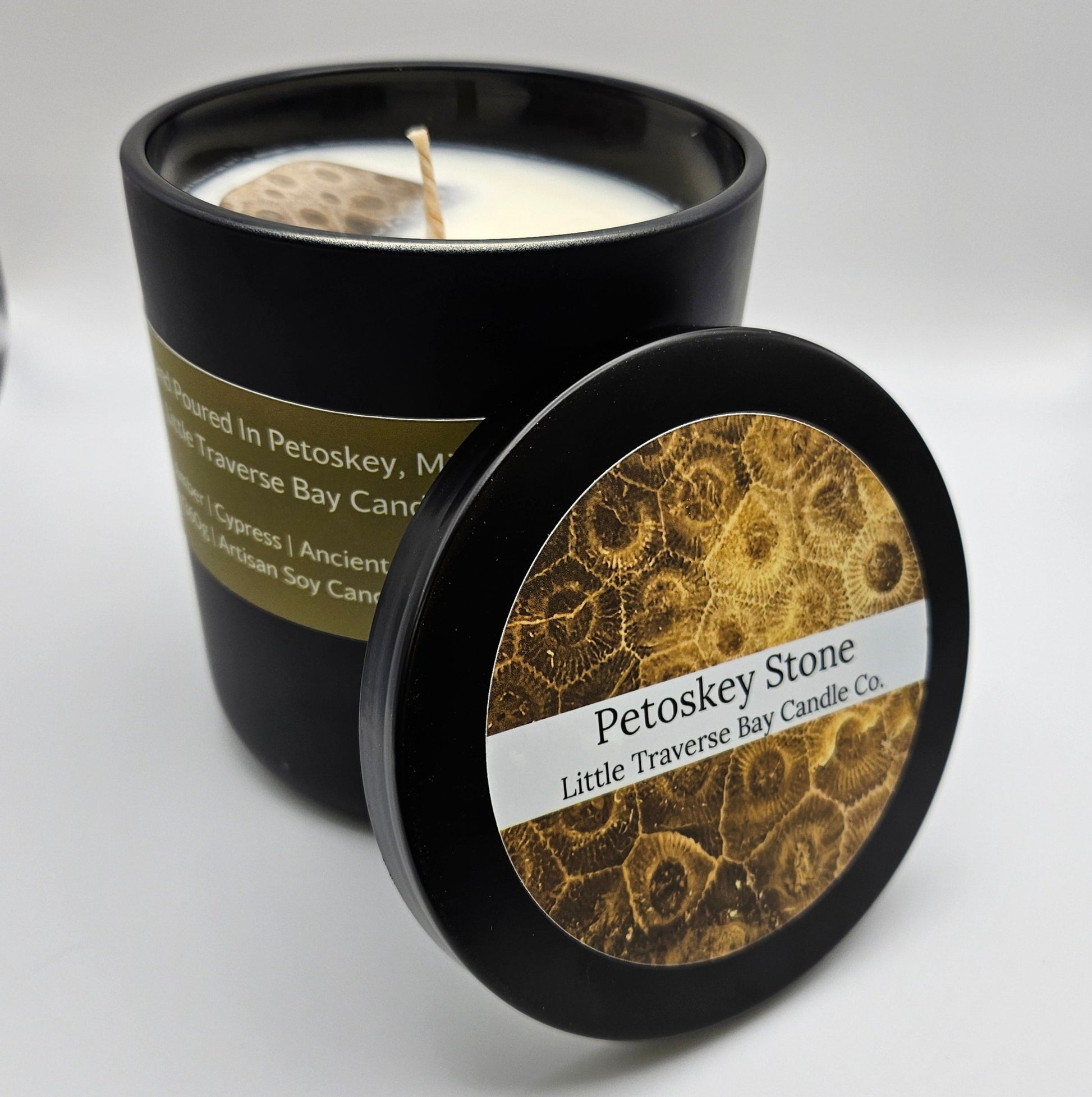 Close up image of the large 12.5 ounce candle with the lid off and leaning up against it with the lid label showing outward. It says Petoskey Stone Little Traverse Bay Candle Co. The inside of the candle shows a sizely petoskey stone with a lovely pattern embedded into the top of the wax with just a bit peeking out. There is a cotton wick in the center, ready for burning.