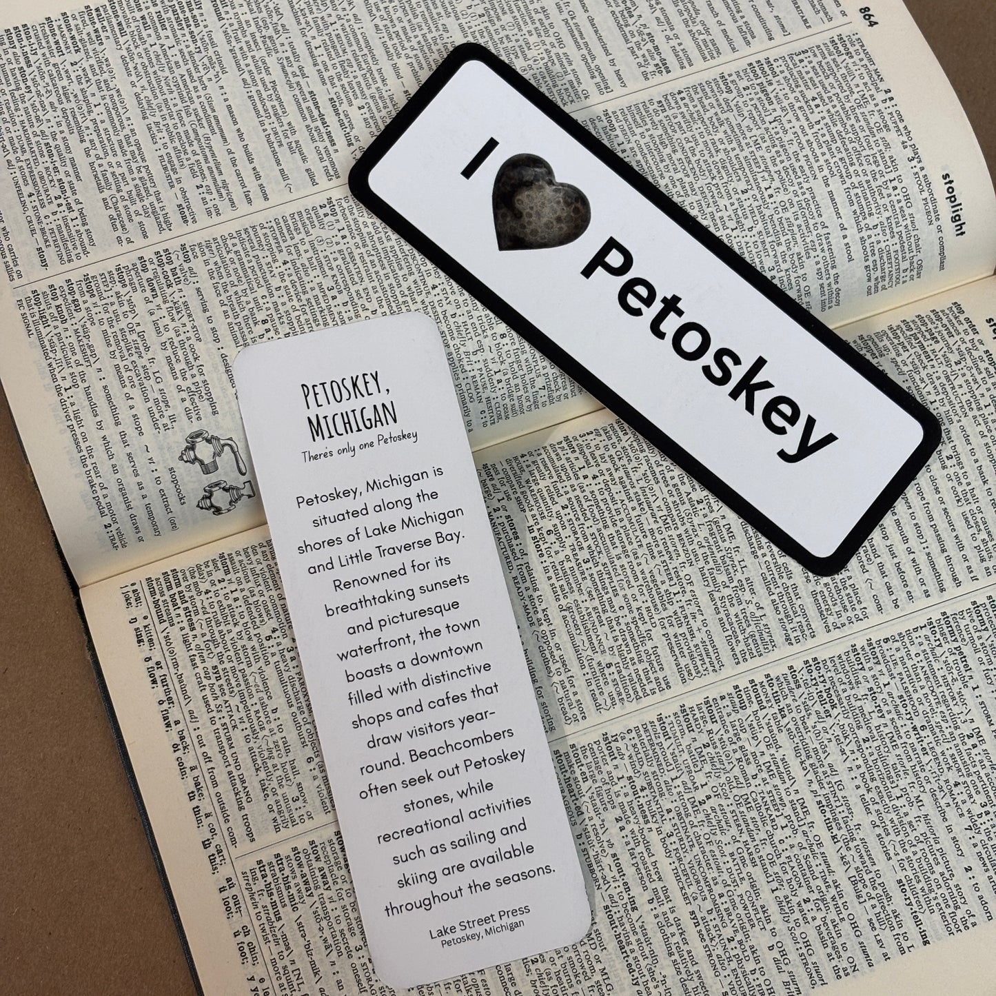 Two printed bookmarks placed on an open book: one rectangular white tag with a black border and the text “I ❤️ Petoskey,” and one vertical white tag with descriptive text about Petoskey, Michigan.