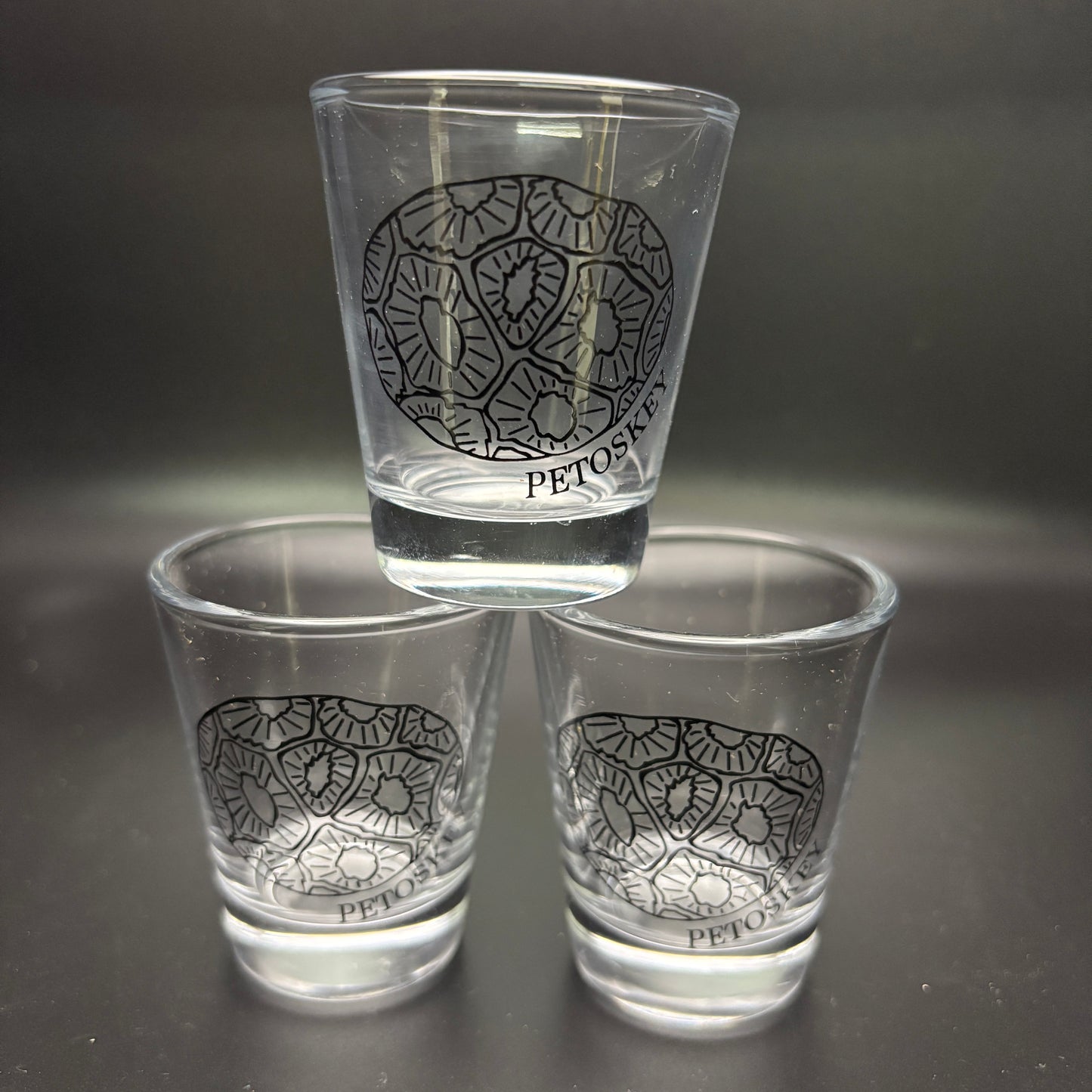 Three clear shot glasses with a black petoskey stone design on a dark background