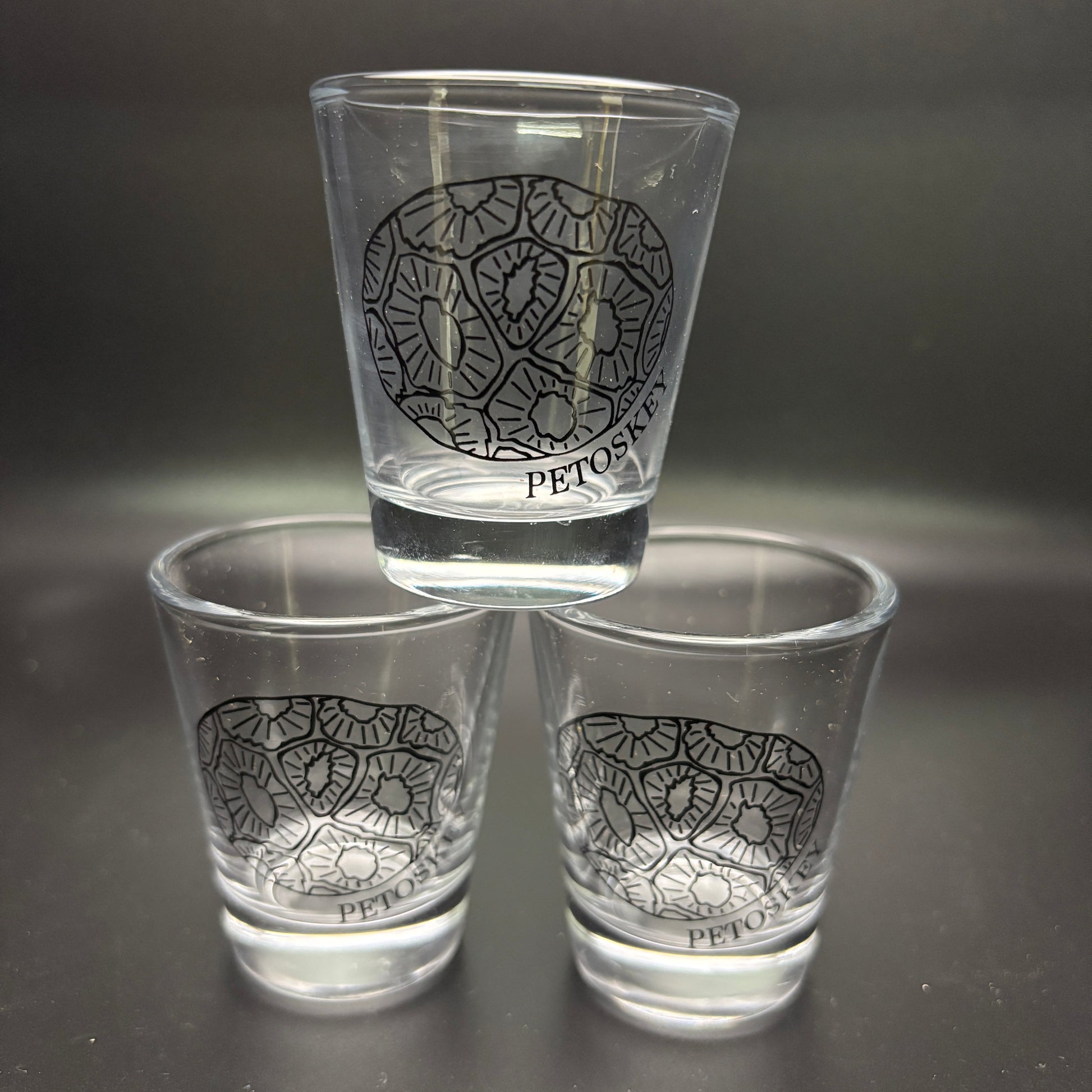Three clear shot glasses with a black petoskey stone design on a dark background