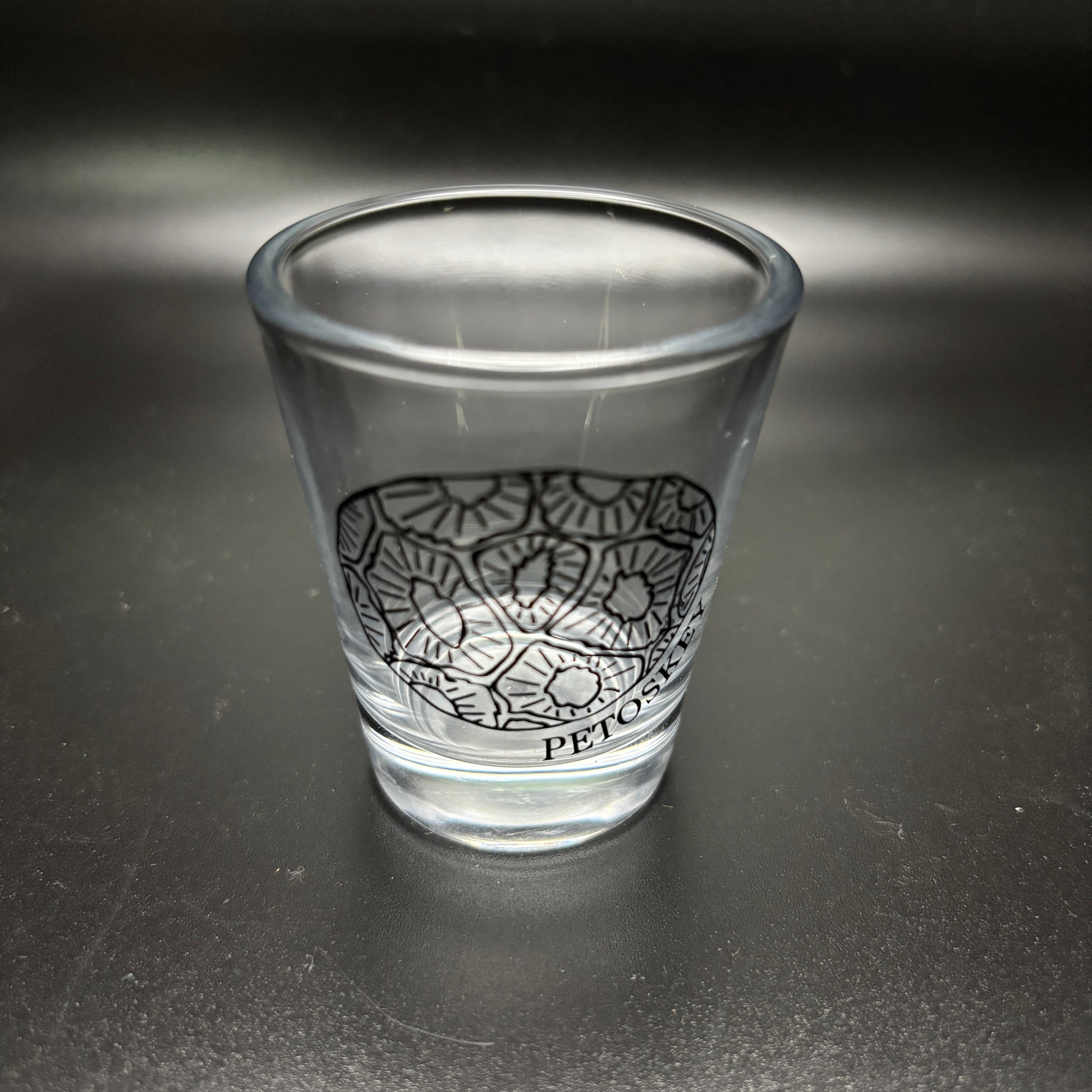 Clear glass shot glass with black design of a petoskey stone and text "Petoskey" on a dark surface