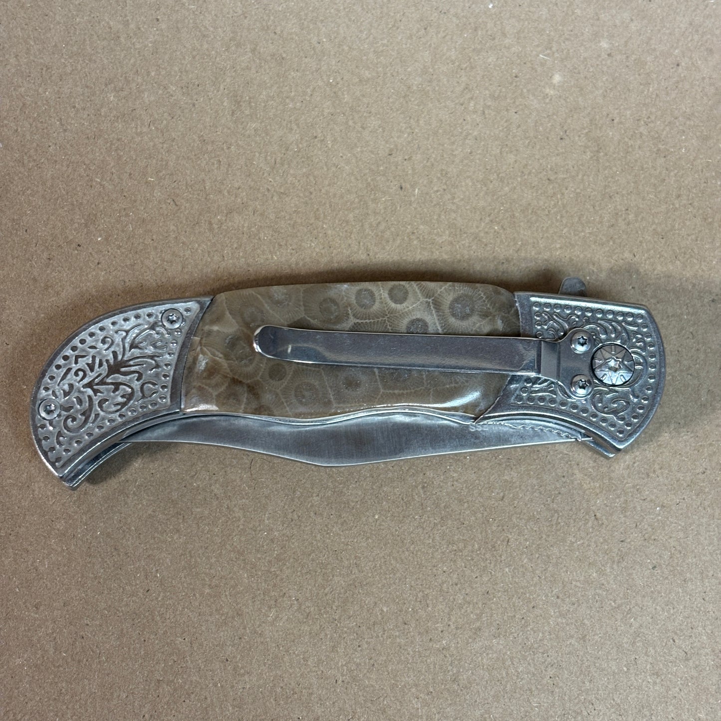 Folding knife with decorative Petoskey stone handle showing the back with a belt clip on a beige background