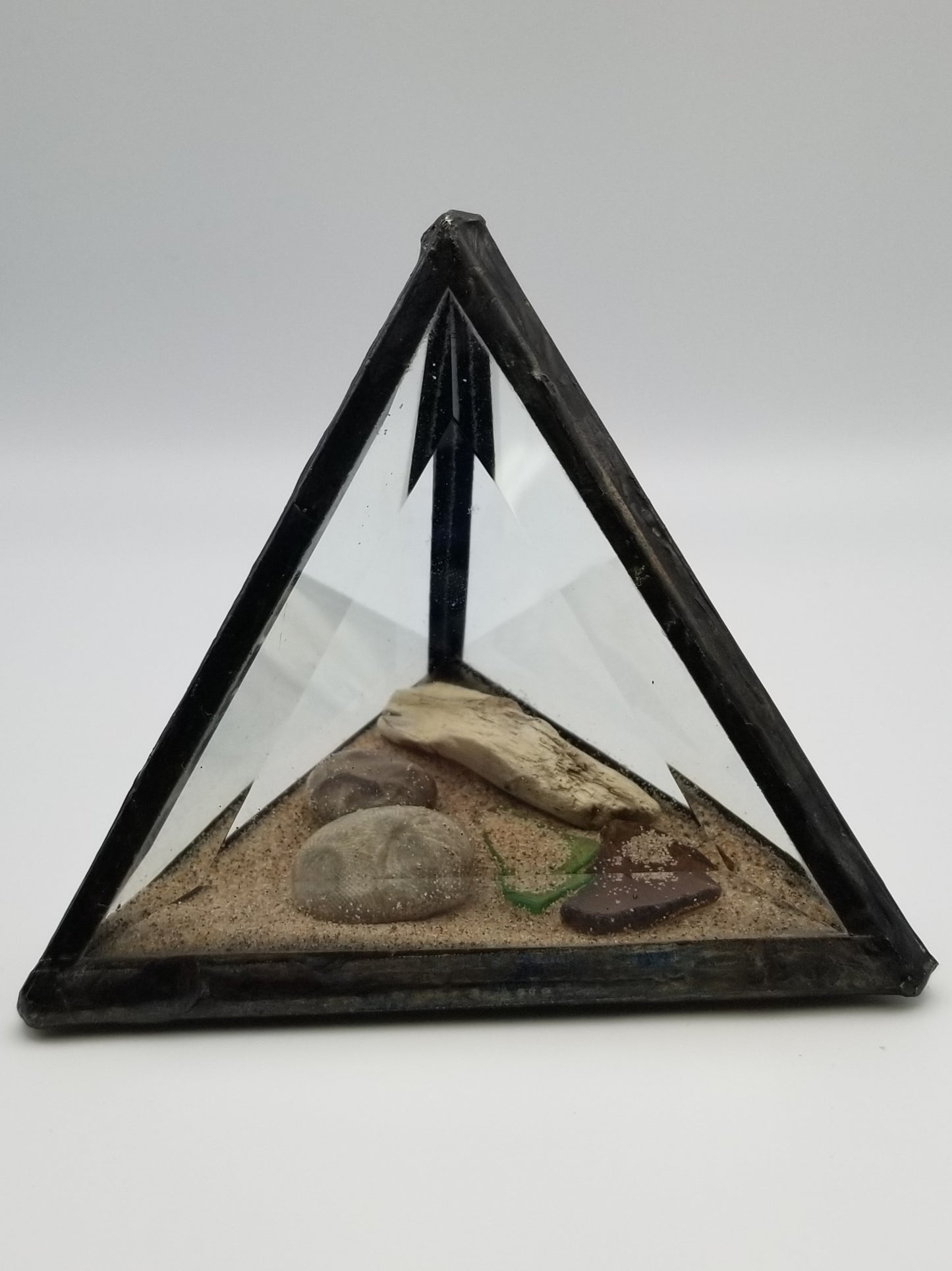 Side view of a fully enclosed glass triangle. Inside there is a thin layer of sand, one petoskey stone, two pieces of driftwood, and two pieces of beach glass.