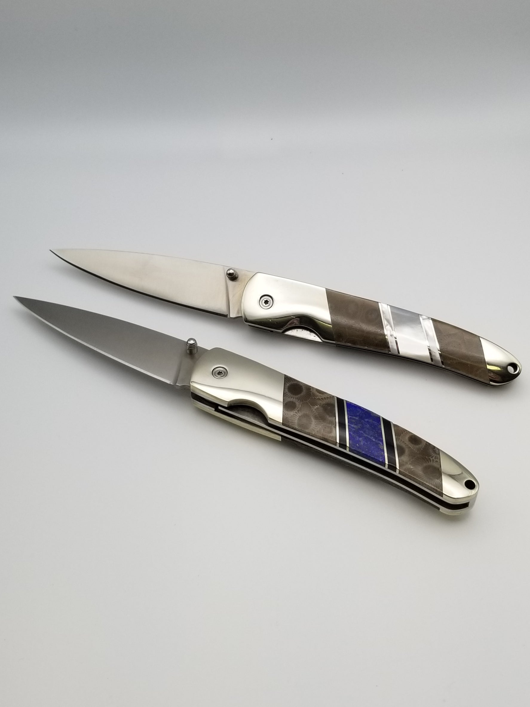 Two assisted open lockback style pocket knives laying opened horizontally. The top one has both petoskey stone and an inlay of mother of pearl. The bottom knife has both petoskey stone and lapis-lazuli inlay.
