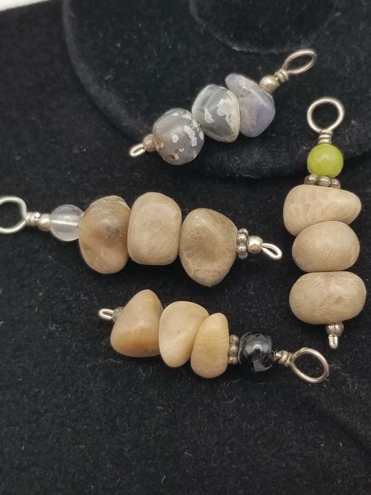 Close up of four (4) dainty stacked stone charms. All have three (3) stacked pebble beads. Three (3) are petoskey stones topped with a clear, green, or black semi-precious round gem bead. The last charm is three (3) stacked mini leland blue pebble beads with no extra bead on top.