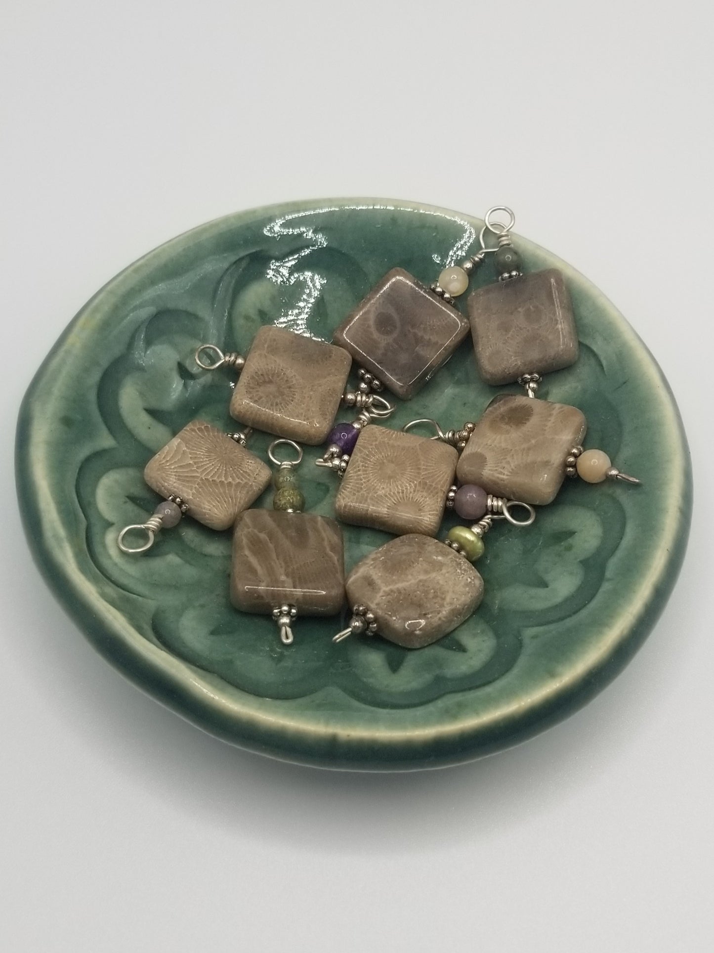 Eight (8) petoskey stone charms in a small, round, dark green dish. Each stone is roughly 1/2 (half) inch or smaller and shaped into a square. There is a single bead atop the charms in an assortment of colors.