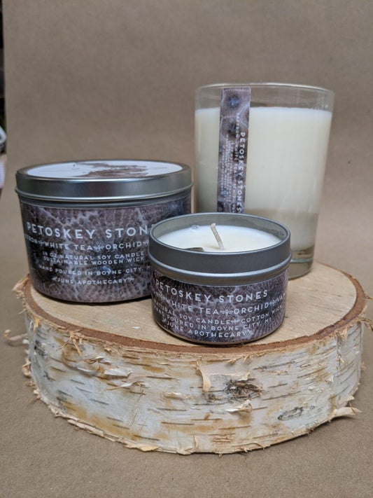 Three candles sitting atop a round slice of birch tree. One candle is a size 4 oz, another is a size 12 oz, and the other is a large glass jar. All three candles feature a petoskey stone embedded on top of the wax.