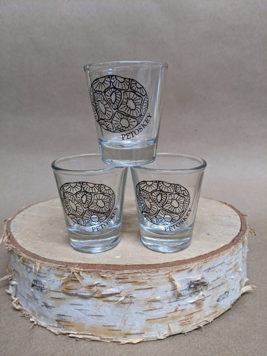 Three (3) shot glasses stacked into a small pyramid atop a round piece of cut birch tree. A locally designed logo is on them with a hand-drawn petoskey stone and "petoskey" written out under it.
