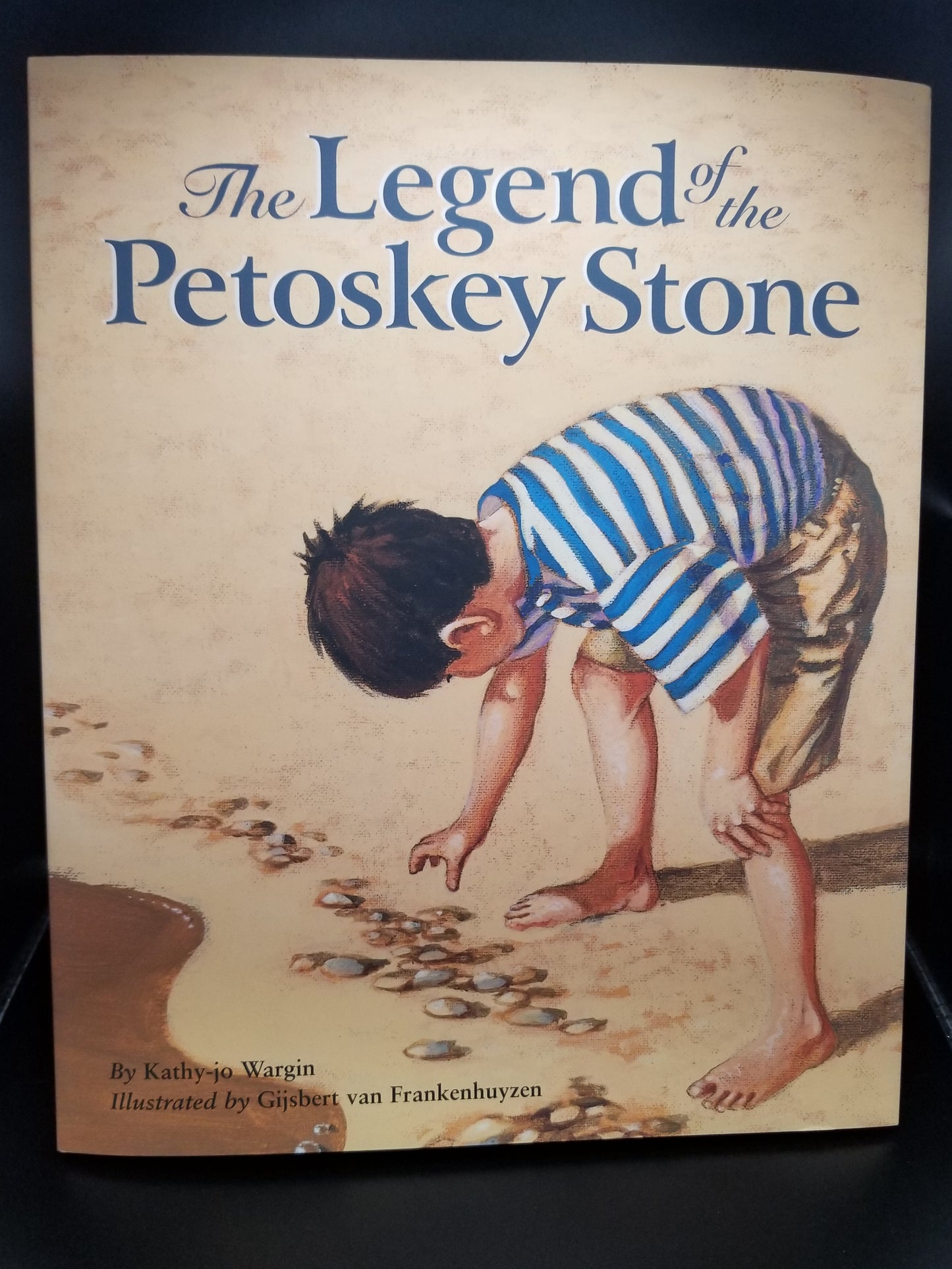 Image of the front cover of the children's book: The Legend of the Petoskey Stone by Kathy jo Wargin. Cover shows a small boy leaning over to pick up a stone on a rock-lined beach shoreline.