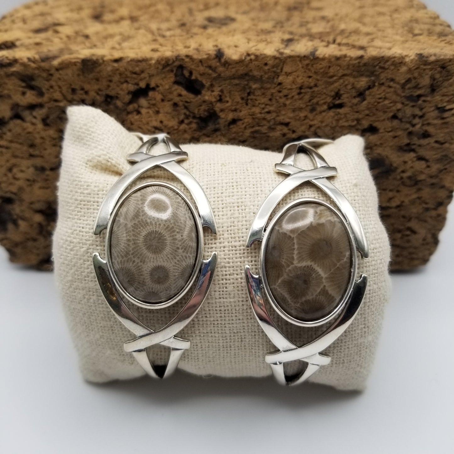 Two .925 sterling silver semi-flexible cuffs featuring a crossed arches design on either side of the bezel. The left cuff has a cab made from a light grey/brown petoskey stone and the right cuff's petoskey stone cab is a darker grey/brown.