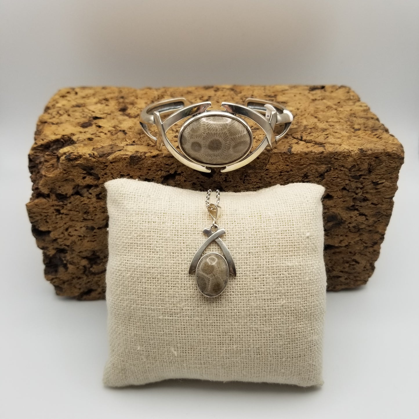 The elegant sterling silver crossed arches necklace with rounded bezel set petoskey stone sits on a canvas mini pillow below a sterling silver cuff bracelet featuring an intricate and classy arch design in silver on either side of a large rounded and polished petoskey stone that has been bezel set. Made in Michigan.