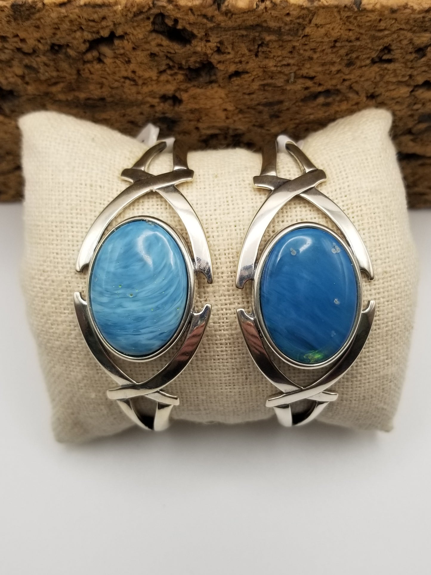 Two .925 sterling silver semi-flexible cuffs featuring a crossed arches design on either side of the bezel. The left cab has a light blue stone cab and the right cuff has a medium blue cab set in the bezel.