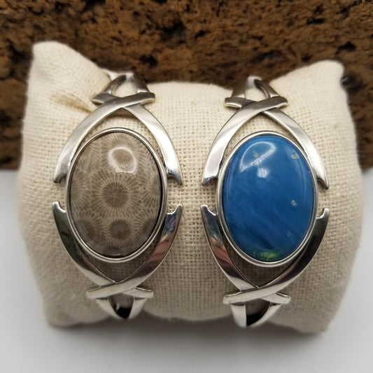 Two .925 sterling silver semi-flexible cuffs featuring a crossed arches design on either side of the bezel. A petoskey stone cab is in the left cuff and a leland blue in a medium blue hue is on the right.