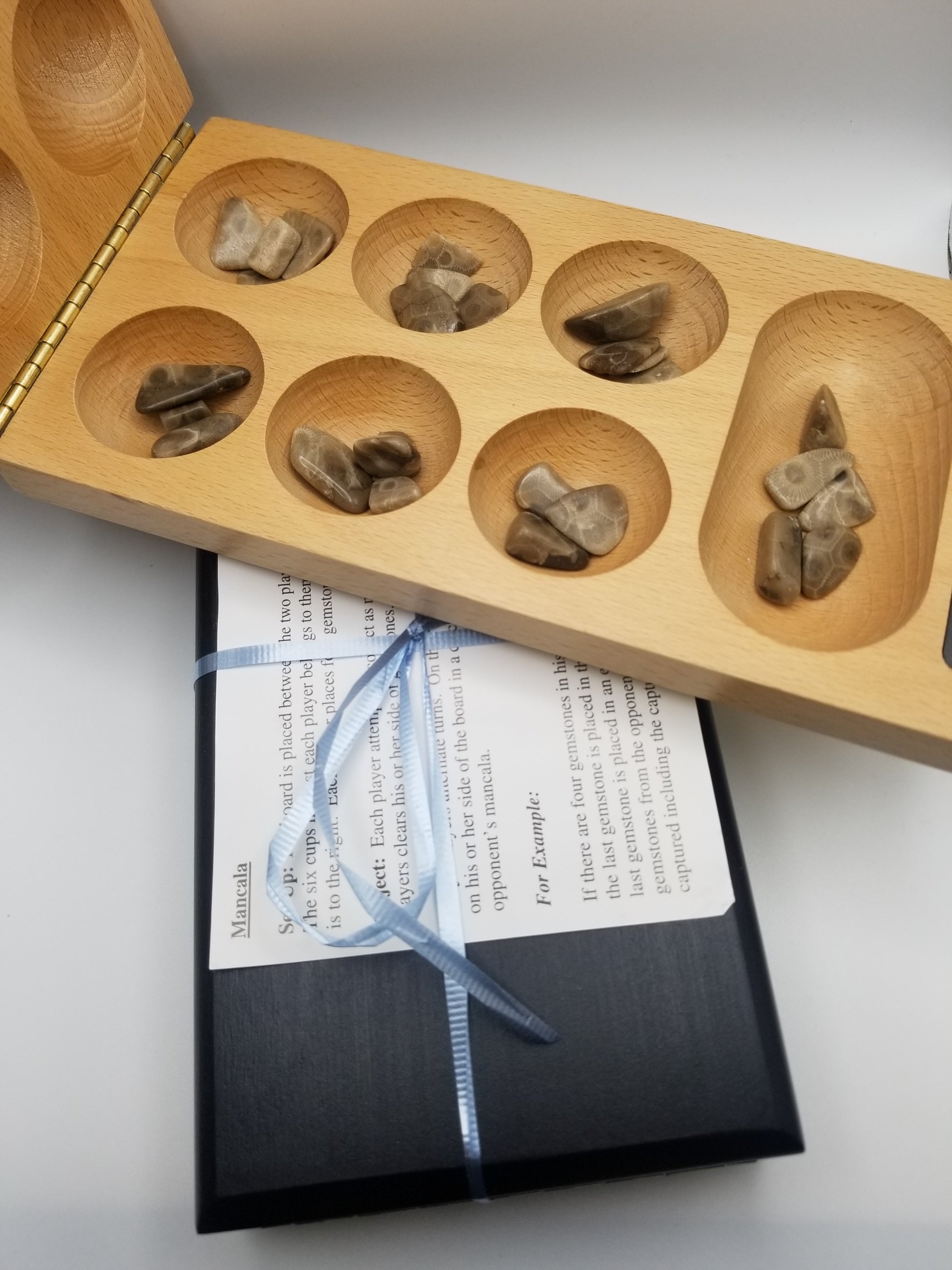 closed up black mancala board with a light blue ribbon wrapped around it and mancala game instructions on top. On top of that is an opened tan mancala board showing off the petoskey stone pebble game pieces in each indented space.