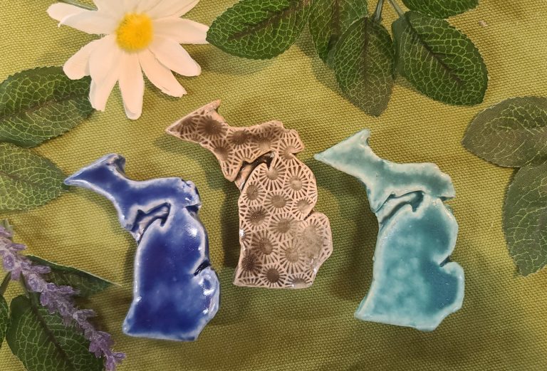 Three handmade clay magnets facing forward on a piece of green fabric. They are shaped with upper and lower Michigan connected as a single magnet. From left to right they are: dark blue, petoskey stone pattern in grey, and light blue.