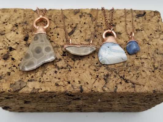 Close up of four necklace charms with the necklace chains hang back from the top. Sitting on top of a cork block. From left to right there is a large petoskey stone with a single bail, a small horizontal petoskey stone with double bails, a large Leland Blue with a single bail, and a small leland blue with a single bail. The Lelands have a wide color variety, with the two shown being opposite spectrums of dark and light. 
