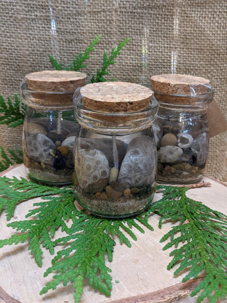 Three glass rounded jars lined up on a burlap fabric with spruce leaves. They all have multiple petoskey stones and beach pebbles and sand set in clear gel candle with a cotton wick.