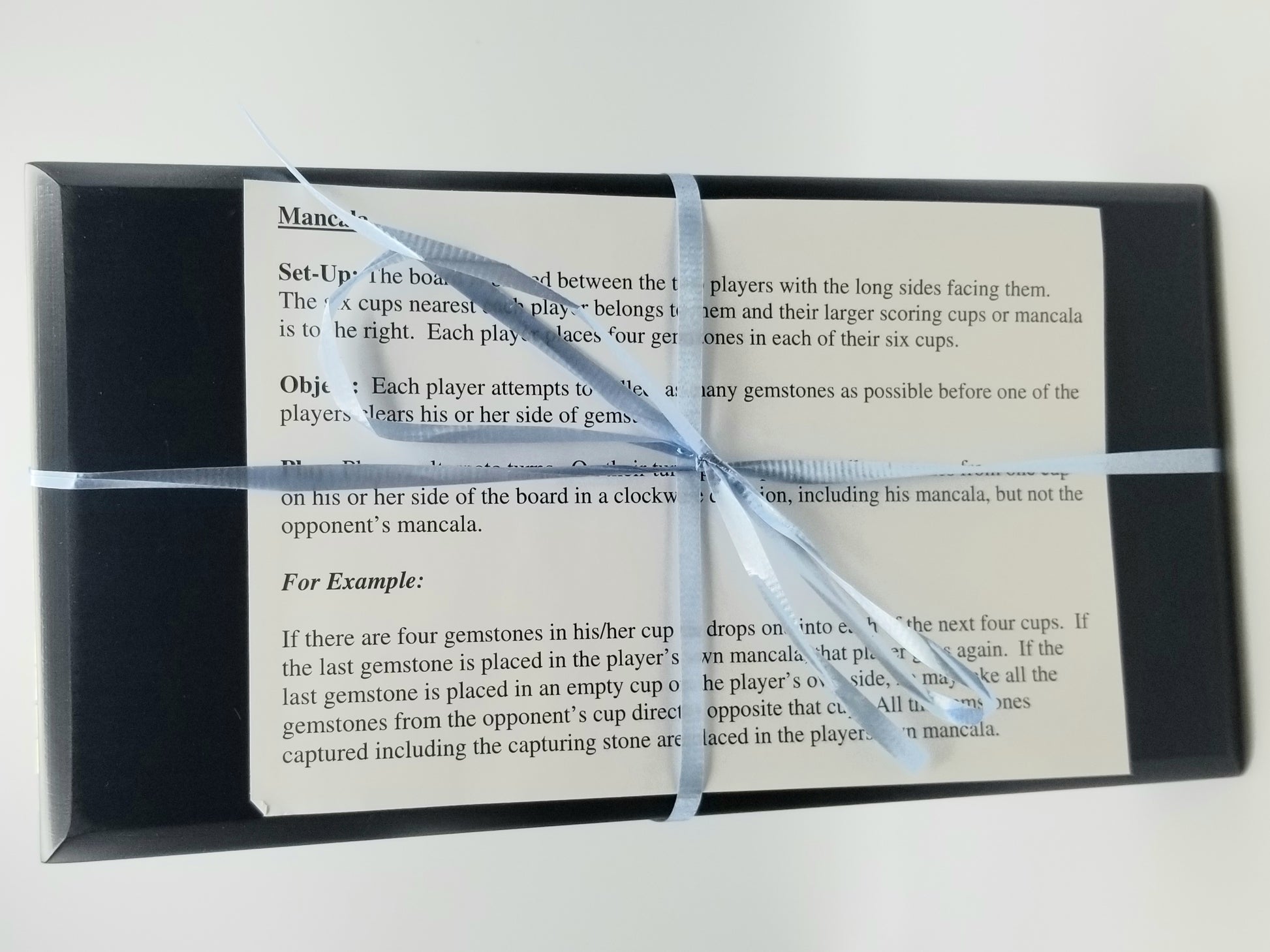 Closed up image of our black mancala board wrapped up in a thin light blue ribbon with the game's instructions tucked into the ribbon.